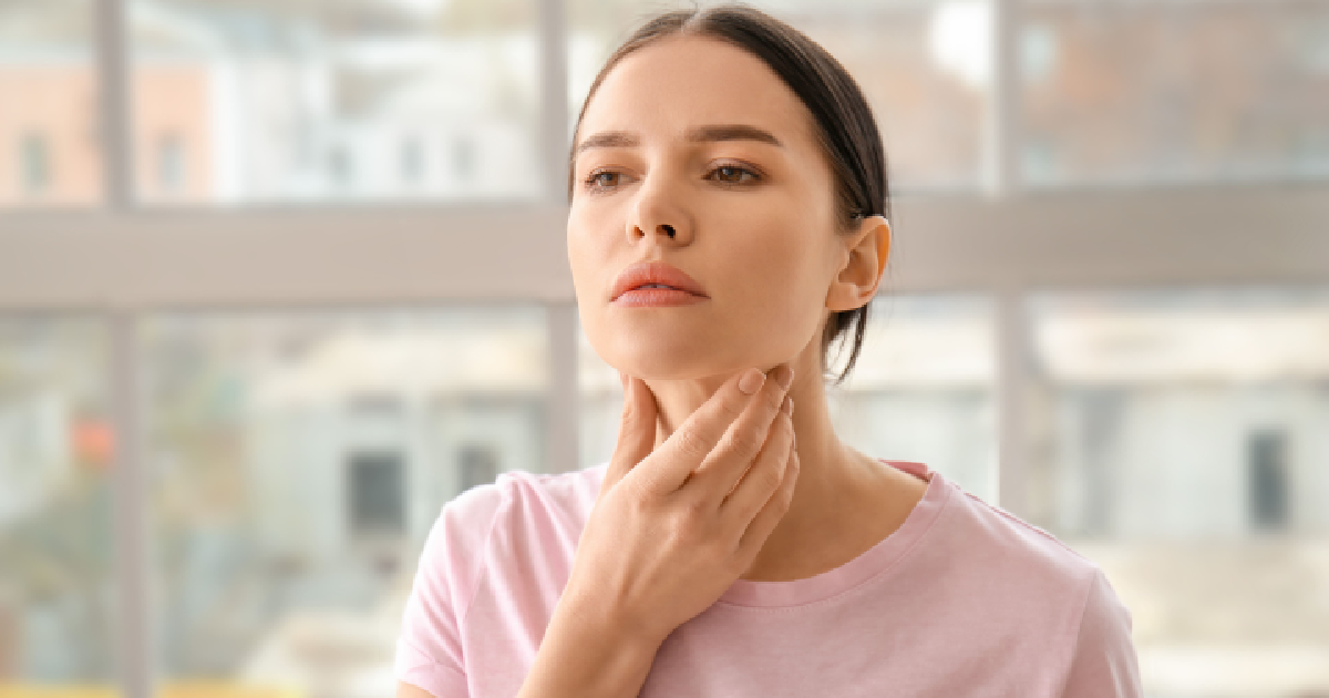 Young woman touching her neck, appearing concerned, possibly checking for thyroid issues related to Thyroid Hormone Therapy; standing indoors in front of large windows in Powell, WY.