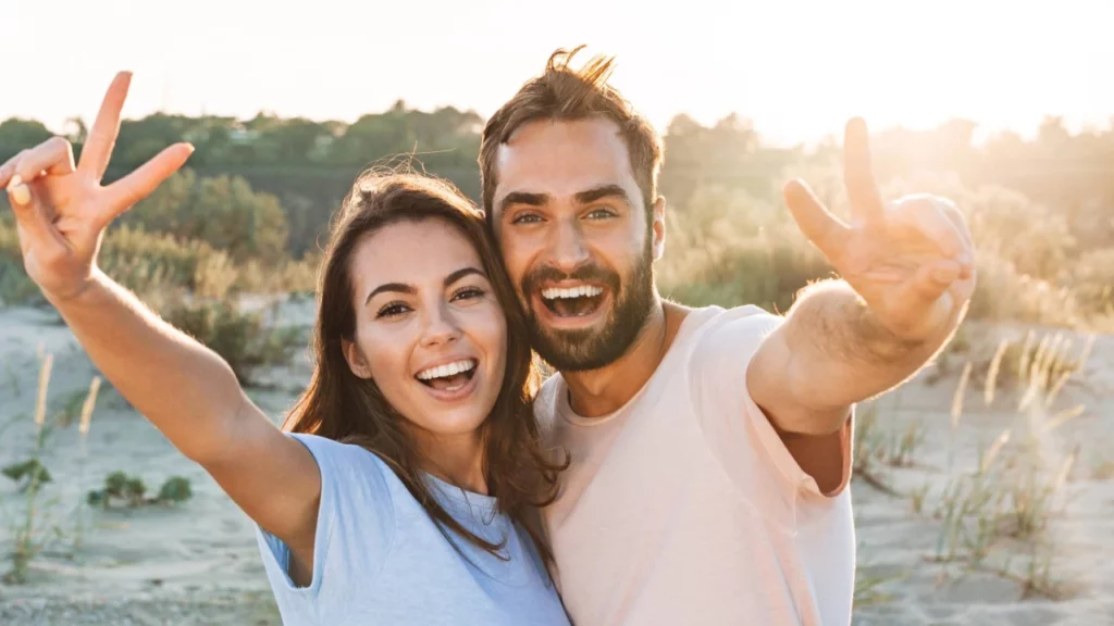 Smiling couple flashes peace signs at the beach, illustrating well-being and vitality after BHRT in Powell, WY.